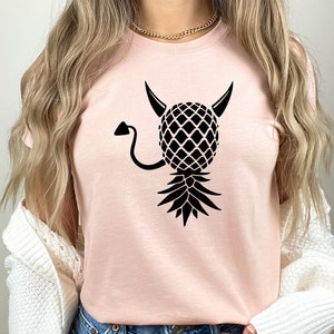 May include: A pink t-shirt with a black pineapple design. The pineapple has horns and a tail, giving it a devilish appearance.