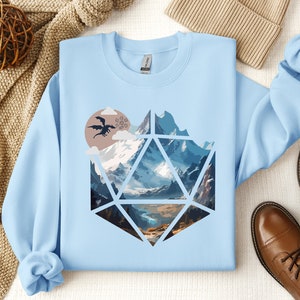 Pode incluir: Camisola azul claro com um gr&aacute;fico de uma cordilheira dentro de um dado azul e branco, com um drag&atilde;o voando no c&eacute;u acima das montanhas.