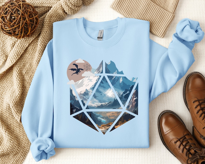 Pode incluir: Camisola azul claro com um gr&aacute;fico de uma cordilheira dentro de um dado azul e branco, com um drag&atilde;o voando no c&eacute;u acima das montanhas.