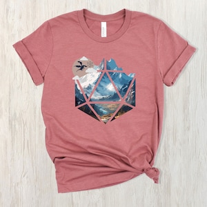Pode incluir: Uma camiseta marrom-avermelhada com um gr&aacute;fico de uma paisagem montanhosa dentro de uma forma geom&eacute;trica que se assemelha a um dado de vinte lados. Um drag&atilde;o preto voa no c&eacute;u acima das montanhas.