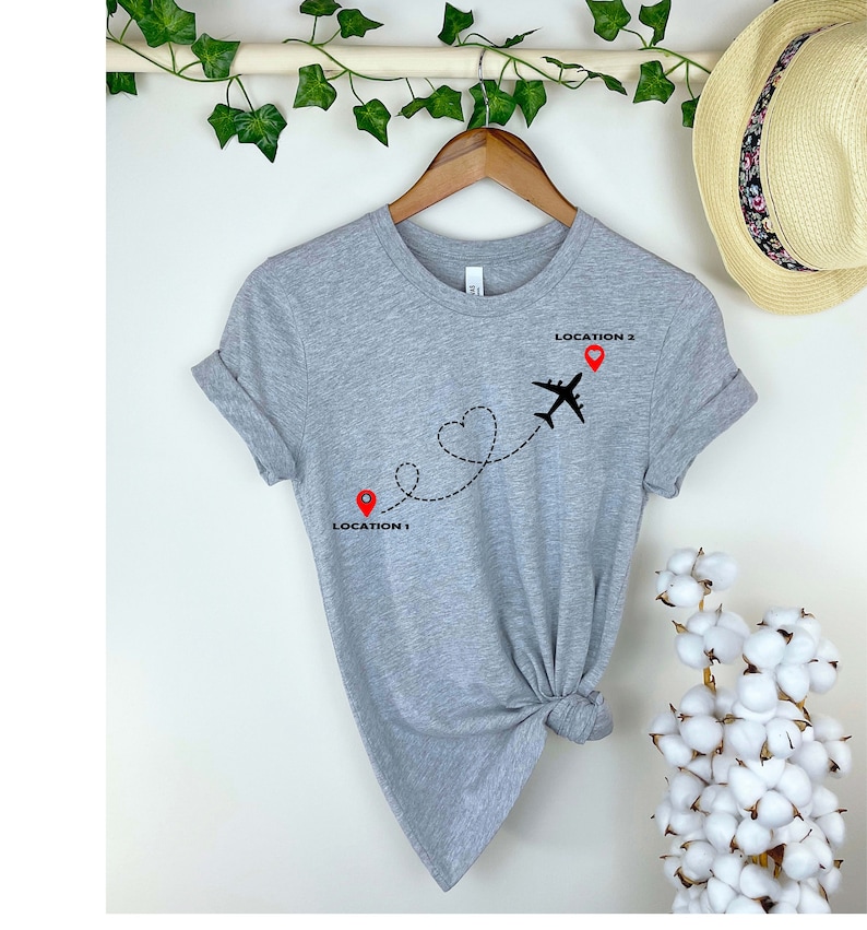Puede incluir: Camiseta gris con un avi&oacute;n negro volando sobre una l&iacute;nea en forma de coraz&oacute;n. La l&iacute;nea conecta dos alfileres rojos etiquetados como "LOCATION 1" y "LOCATION 2".