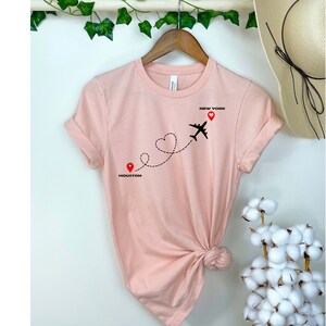 Puede incluir: Una camiseta rosa con un gr&aacute;fico de avi&oacute;n negro volando de Houston a Nueva York. La ruta de vuelo es una l&iacute;nea punteada con una forma de coraz&oacute;n en el medio. Las ciudades est&aacute;n etiquetadas con alfileres rojos.