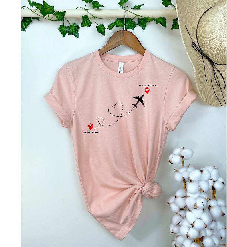 Puede incluir: Una camiseta rosa con un gr&aacute;fico de avi&oacute;n negro volando de Houston a Nueva York. La ruta de vuelo es una l&iacute;nea punteada con una forma de coraz&oacute;n en el medio. Las ciudades est&aacute;n etiquetadas con alfileres rojos.