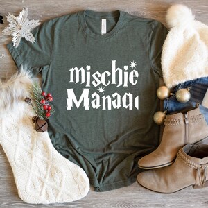 May include: A green t-shirt with the text "mischie* Manaqi" in white. The shirt is laying on a wooden surface with a white knitted stocking, a white fuzzy hat, a pair of brown suede ankle boots, a pair of blue jeans, and a white mug.