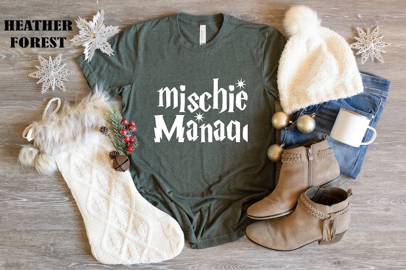 May include: A green t-shirt with the text "mischie* Manaqi" in white. The shirt is laying on a wooden surface with a white knitted stocking, a white fuzzy hat, a pair of brown suede ankle boots, a pair of blue jeans, and a white mug.