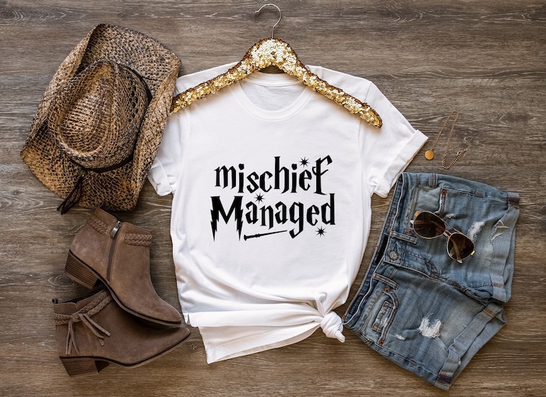 May include: White t-shirt with black text that reads "mischief managed" with a wand and stars. The shirt is styled with a brown cowboy hat, brown boots, and denim shorts.