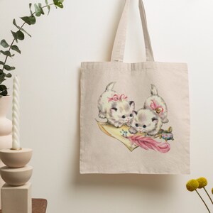 May include: A white canvas tote bag with a vintage illustration of three kittens wearing pink bows and playing with a feather pen and inkwell. The kittens are on a pink and yellow piece of paper.