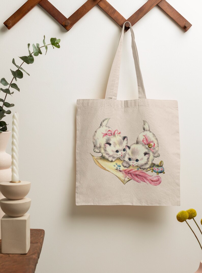 May include: A white canvas tote bag with a vintage illustration of three kittens wearing pink bows and playing with a feather pen and inkwell. The kittens are on a pink and yellow piece of paper.