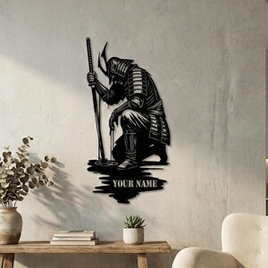 May include: Black metal wall art depicting a kneeling samurai warrior holding a katana. The artwork includes the text "YOUR NAME" at the base. The samurai is wearing traditional armor and a helmet. The art is mounted on a light-colored wall.