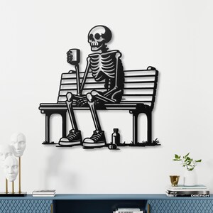 Puede incluir: Escultura de pared de metal negro de un esqueleto sentado en un banco sosteniendo una taza. La escultura está montada en una pared blanca sobre un gabinete azul con libros en la parte superior.