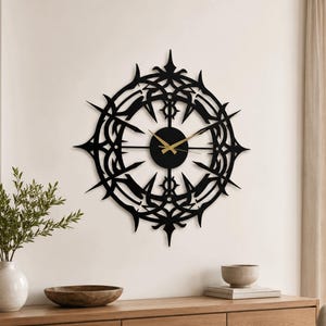 Puede incluir: Un reloj de pared circular con un marco de metal negro y un patrón geométrico complejo, simétrico y en forma de estrella. La esfera tiene un centro negro sólido con manecillas finas de color dorado que ofrecen un contraste marcado.