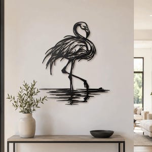 Puede incluir: Una pieza de arte mural de metal negro que representa un flamenco de pie en el agua. El diseño presenta recortes intrincados que definen las plumas del ave y su reflejo en la superficie del agua, creando una silueta detallada en la pared.