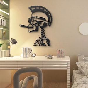 May include: Black metal wall art of a skeleton with a mohawk, sunglasses, and a cigarette hanging from its mouth. The skeleton is wearing a helmet and has a ribcage visible. The art is mounted on a beige wall above a white desk with a lamp, books, and a toy helicopter.