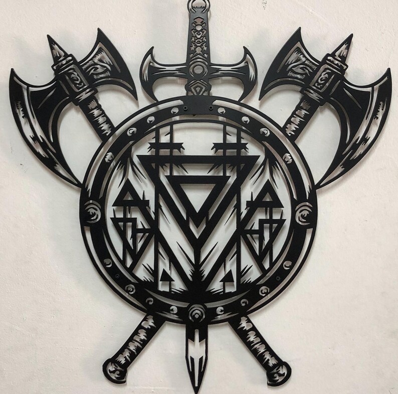 May include: A black metal wall art piece featuring a central circular shield with geometric triangle patterns, flanked by two crossed battle axes and a vertical sword positioned behind the shield, creating a symmetrical weapon-themed design.