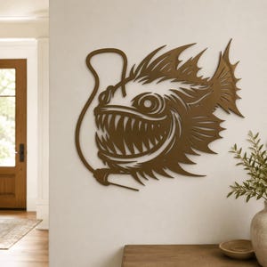 May include: A bronze-colored metal wall art piece shaped like an anglerfish with sharp, pointed teeth, large circular eyes, and a prominent dorsal fin. The fish features a long, curved lure extending from its head, creating a detailed silhouette.