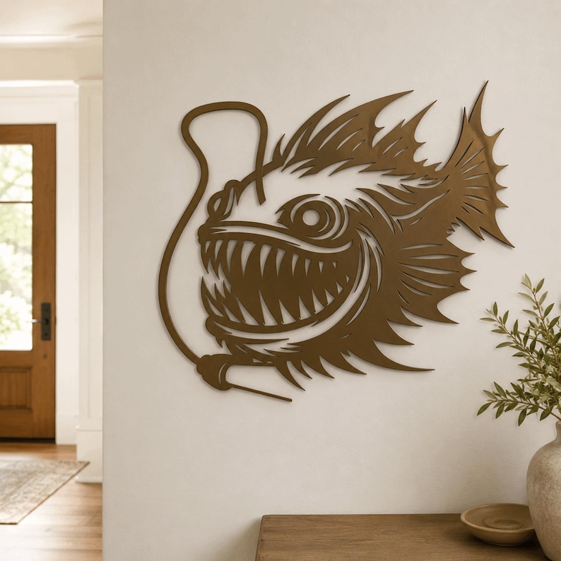 May include: A bronze-colored metal wall art piece shaped like an anglerfish with sharp, pointed teeth, large circular eyes, and a prominent dorsal fin. The fish features a long, curved lure extending from its head, creating a detailed silhouette.