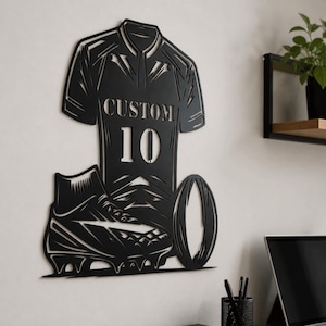 Puede incluir: Una pieza de arte mural de metal negro que muestra una camiseta deportiva con el texto CUSTOM y el número 10, acompañada de una bota de fútbol y un balón de rugby. El diseño está cortado en una lámina de metal con líneas intrincadas.