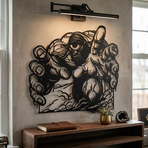 May include: Black metal wall art depicting a cyclops reaching out. The artwork is mounted on a light gray wall, with a black light fixture above. A wooden console table below holds a leather-bound book and a small potted plant.