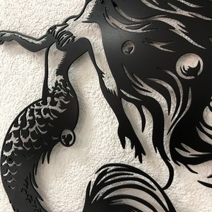 Puede incluir: Una pieza de arte mural de metal negro que presenta la silueta de una sirena con cabello largo y fluido y una cola detallada. El dise&ntilde;o est&aacute; cortado de una sola hoja de metal, mostrando curvas intrincadas y espacios negativos definidos.