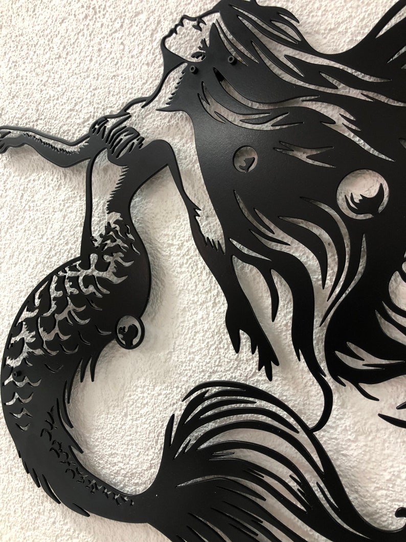 Puede incluir: Una pieza de arte mural de metal negro que presenta la silueta de una sirena con cabello largo y fluido y una cola detallada. El dise&ntilde;o est&aacute; cortado de una sola hoja de metal, mostrando curvas intrincadas y espacios negativos definidos.