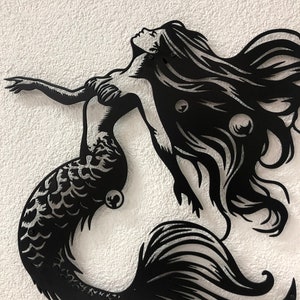 Puede incluir: Una pieza de arte mural de metal negro que presenta la silueta de una sirena con cabello largo y fluido, espalda arqueada y un brazo extendido. El dise&ntilde;o incluye recortes intrincados que representan escamas en la cola y burbujas.