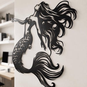 Puede incluir: Una pieza de arte mural de metal negro que presenta la silueta de una sirena con cabello largo y fluido y una cola detallada. El dise&ntilde;o est&aacute; cortado de una sola hoja de material, mostrando patrones intrincados en el cabello y las aletas de la cola.