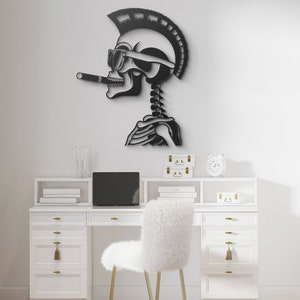 May include: A black metal wall sculpture of a skull with a mohawk, sunglasses, and a cigarette. The skull is in a profile view and is mounted on a white wall. The sculpture is above a white desk with a white chair, a laptop, and other office supplies.