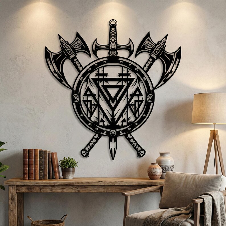 May include: A black metal wall art piece featuring a circular shield design centred behind a vertical sword and two crossed battle axes. The shield displays intricate geometric patterns including triangles and vertical lines within its circular frame.