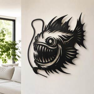 May include: A black metal wall art piece shaped like an anglerfish with a prominent lure, sharp teeth, and spiky fins. The design features intricate cutouts that define the fish's silhouette, eye, and mouth, creating a bold, dark graphic display.