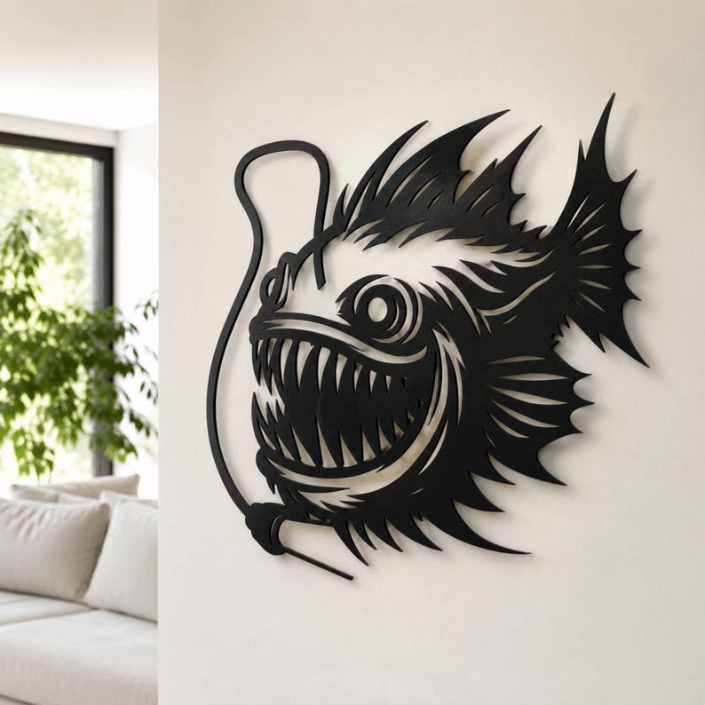 May include: A black metal wall art piece shaped like an anglerfish with a prominent lure, sharp teeth, and spiky fins. The design features intricate cutouts that define the fish's silhouette, eye, and mouth, creating a bold, dark graphic display.
