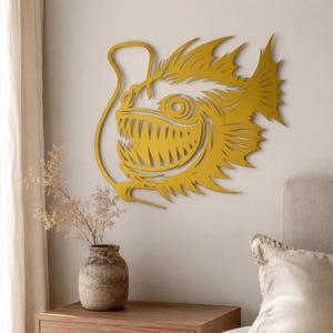 May include: A mustard yellow metal wall art piece shaped like an anglerfish with a prominent lure, sharp teeth, and spiky fins. The design features intricate cutouts that define the fish's eye, mouth, and scales, creating a bold silhouette.