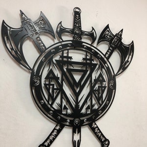 May include: A black metal wall art piece featuring a circular shield design with a central geometric triangle pattern. Two battle axes are positioned behind the shield, and a sword is mounted vertically, extending from the top and bottom.