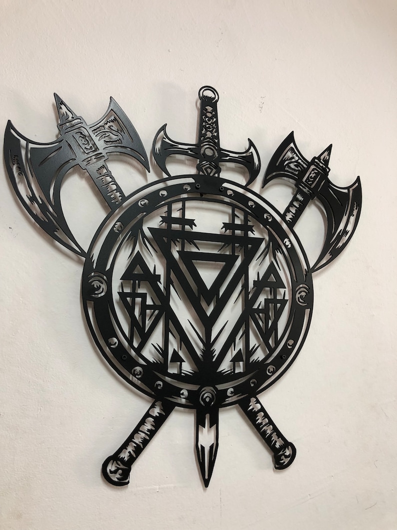 May include: A black metal wall art piece featuring a circular shield design with a central geometric triangle pattern. Two battle axes are positioned behind the shield, and a sword is mounted vertically, extending from the top and bottom.