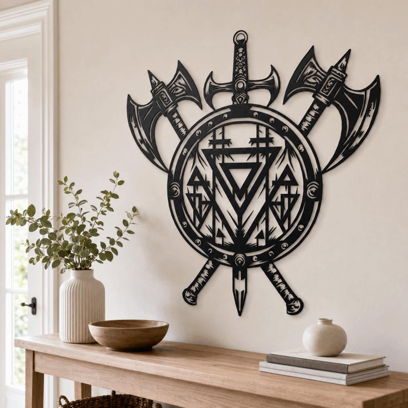 May include: A black metal wall art piece featuring a central circular shield design with a sword pointing downward and two crossed battle axes behind it. The shield contains intricate geometric patterns including triangles and vertical lines.