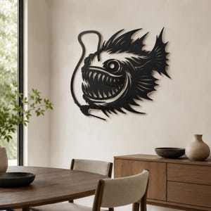 May include: A black metal wall art piece shaped like an anglerfish with sharp, pointed teeth and a long, curved lure extending from its head. The design features intricate cutouts and jagged fins, creating a bold silhouette against the wall.