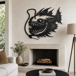 May include: A black metal wall art piece shaped like an anglerfish with a prominent lure, sharp teeth, and spiky fins. The design features a cutout silhouette style with detailed lines defining the fish's scales, eye, and aggressive facial features.