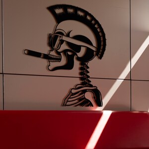 May include: A black and white silhouette of a skull with a mohawk and sunglasses holding a cigarette. The skull is depicted as a skeleton with visible ribs. The image is on a light beige background.