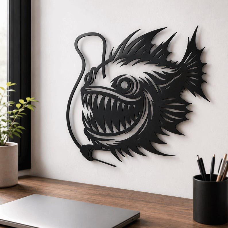 May include: A black metal wall art piece featuring a stylized anglerfish with sharp, pointed teeth, large circular eyes, and a prominent dorsal fin, with a curved line extending from its head to represent a fishing lure.