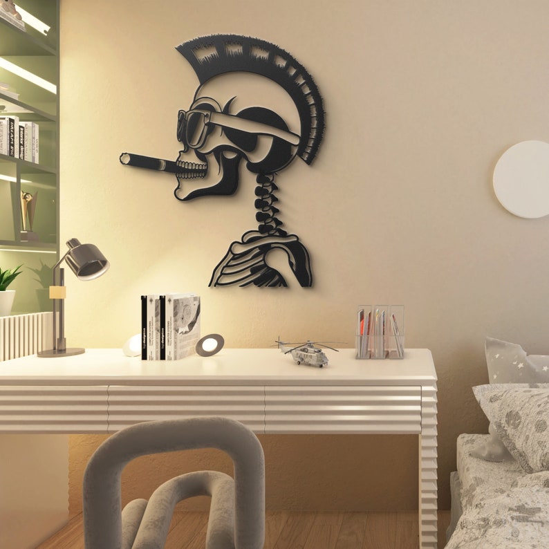 May include: Black metal wall art of a skeleton with a mohawk, sunglasses, and a cigarette hanging from its mouth. The skeleton is wearing a helmet and has a ribcage visible. The art is mounted on a beige wall above a white desk with a lamp, books, and a toy helicopter.