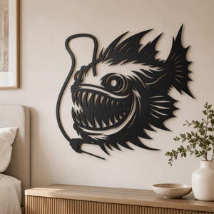 May include: A black, flat, laser-cut metal wall art piece depicting an anglerfish with sharp, pointed teeth, large circular eyes, and a prominent dorsal lure. The design features intricate, jagged fins and a curved, slender line extending from the head.