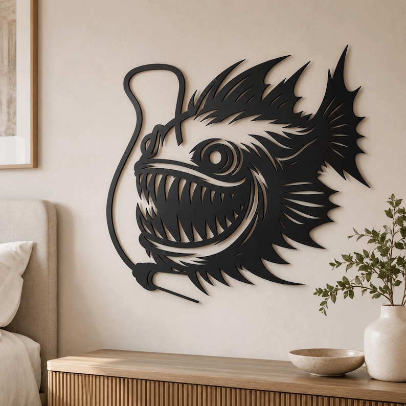 May include: A black, flat, laser-cut metal wall art piece depicting an anglerfish with sharp, pointed teeth, large circular eyes, and a prominent dorsal lure. The design features intricate, jagged fins and a curved, slender line extending from the head.
