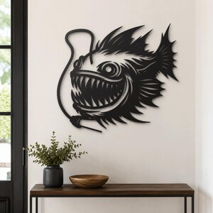 May include: A black metal wall art piece shaped like an anglerfish with a prominent lure, sharp teeth, and spiky fins. The design features intricate cutouts that define the fish's aggressive facial features and scales, mounted on a plain wall.