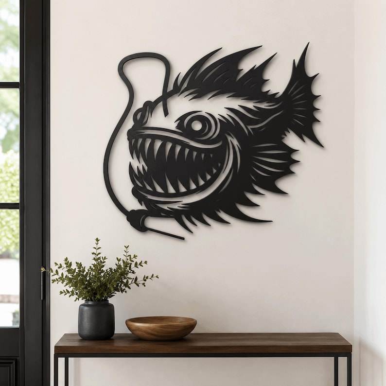 May include: A black metal wall art piece shaped like an anglerfish with a prominent lure, sharp teeth, and spiky fins. The design features intricate cutouts that define the fish's aggressive facial features and scales, mounted on a plain wall.