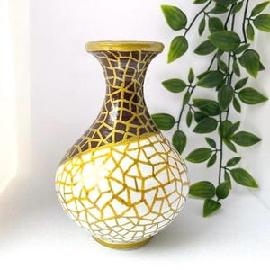 May include: A white ceramic vase with a gold and brown geometric pattern. The vase has a round body and a narrow neck.