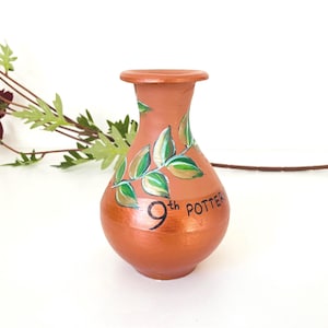 May include: A terracotta-colored vase with hand-painted green and gold leaves. The vase has a rounded body and a narrow neck. The text "9th POTTER" is written on the vase. Decorative floral elements are in the background.