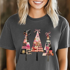 May include: A dark gray t-shirt with a design of three pink, black, and gold patchwork Christmas trees with pink bows. The text "Comfort Colors Pepper" is visible in the bottom left corner of the image.
