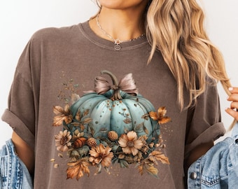 May include: A brown t-shirt with a teal blue pumpkin with fall leaves and flowers. The pumpkin has a brown bow on top.
