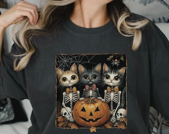 May include: A dark grey t-shirt with a graphic of three black cats holding a jack-o'-lantern. The cats are wearing bow ties and have skeletal features. The graphic is set against a dark background with a spider web and a spider.