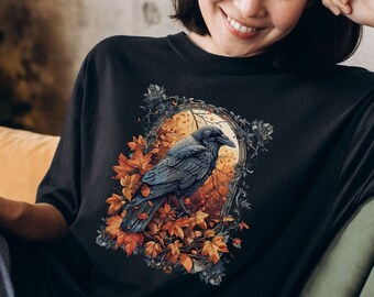 May include: Black t-shirt featuring a detailed illustration of a raven perched among autumn leaves within an ornate frame. The design includes shades of orange, brown, and gray. The text "Comfort Colors Black" is displayed on the bottom right.