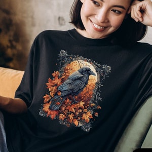 May include: Black t-shirt featuring a detailed illustration of a raven perched among autumn leaves within an ornate frame. The design includes shades of orange, brown, and gray. The text "Comfort Colors Black" is displayed on the bottom right.